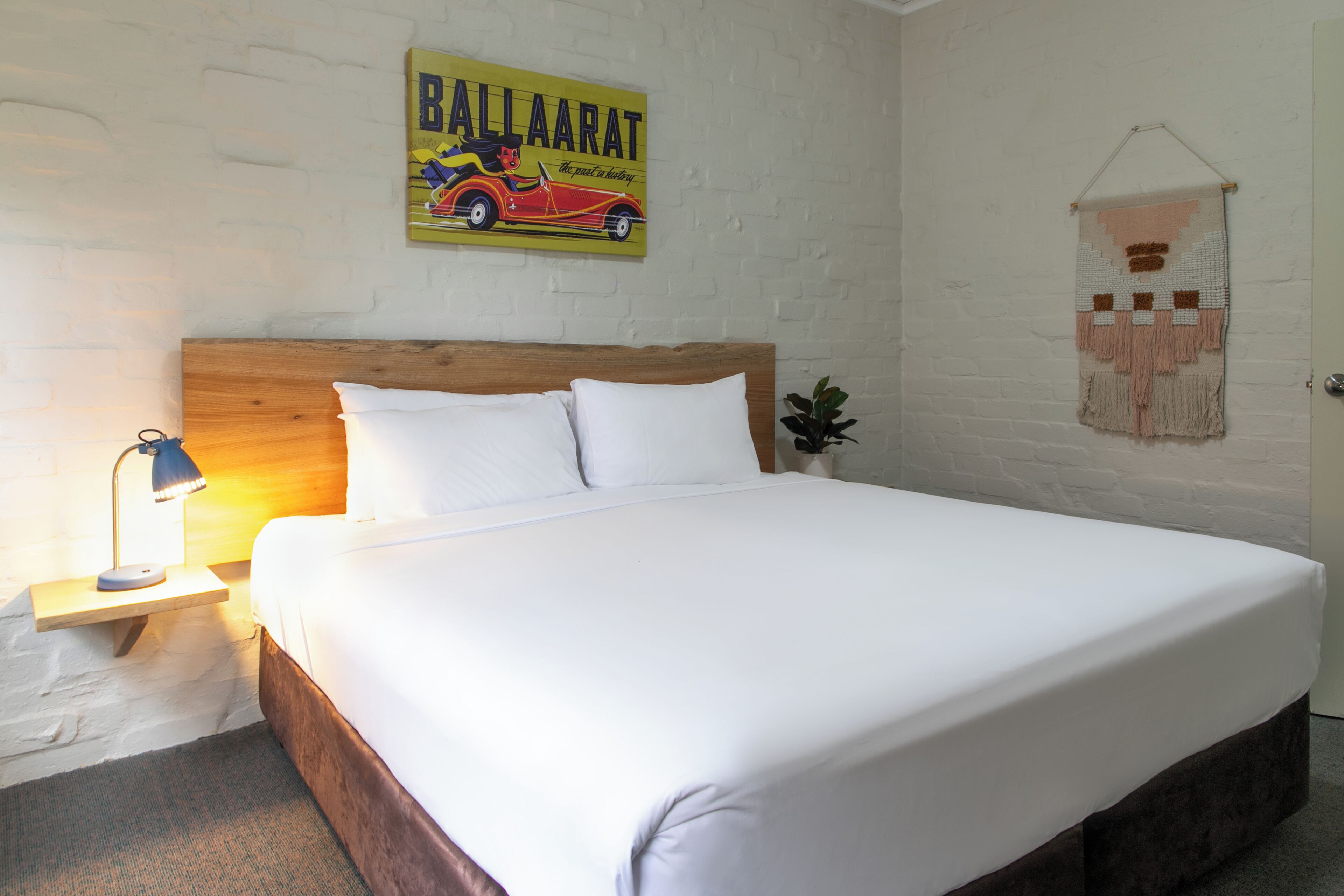 Una cama bien tendida con sábanas blancas en una habitación con pared de ladrillo blanco, cabecera de madera, mesita de noche con lámpara y un adorno colgante.