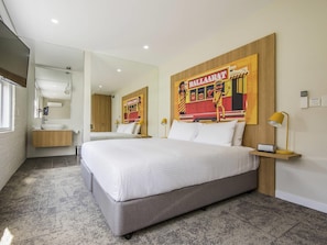 A hotel room with a large bed, a bedside table with a lamp, a mirror, a bathroom with a sink and toilet, and a wall with a colorful poster. - Mercure Ballarat Hotel and Convention Centre (Golden Point (Ballarat))