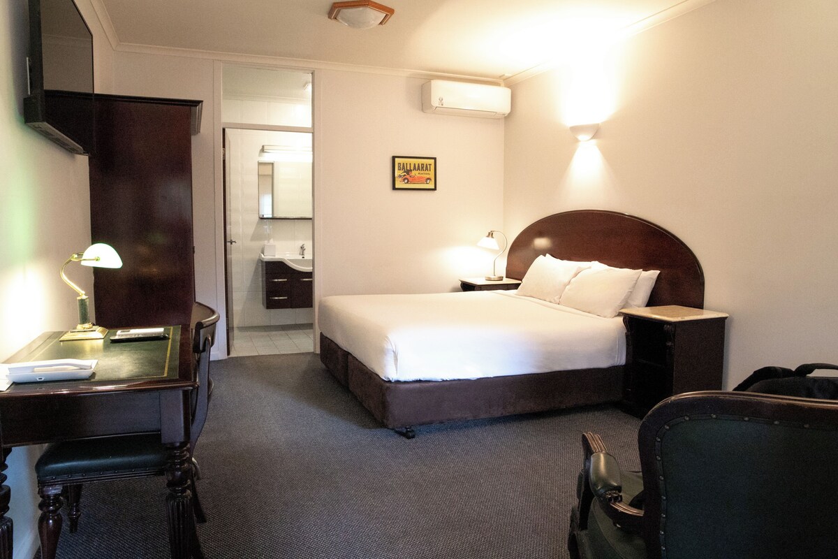 Une chambre d’hôtel avec un lit, des tables de chevet, un bureau avec une lampe, une chaise et une valise.
