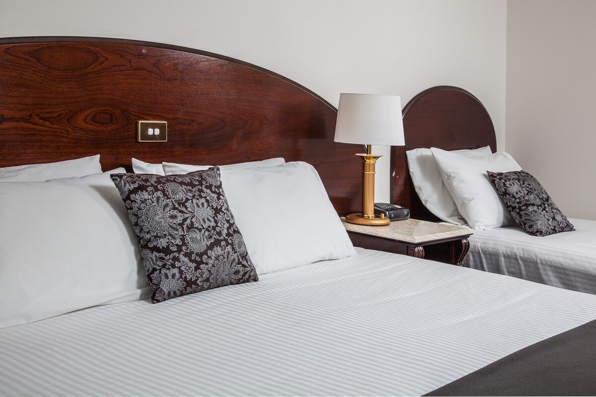 Une chambre d’hôtel avec deux lits, chacun agrémenté de draps blancs et d’oreillers à motifs. On y trouve une table de chevet avec une lampe et un petit plateau.