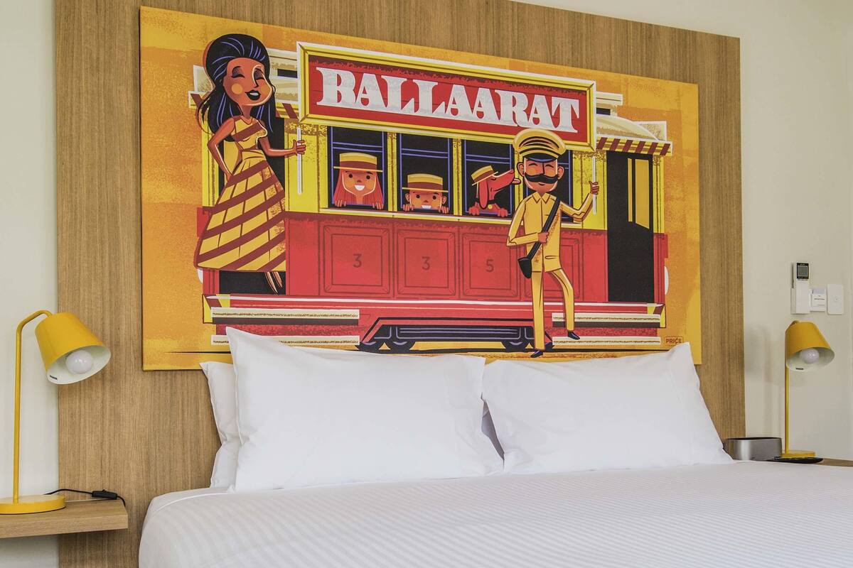 Een hotelkamer met een bed, twee bedlampjes, een wandklok en een kleurrijk schilderij van een tram.