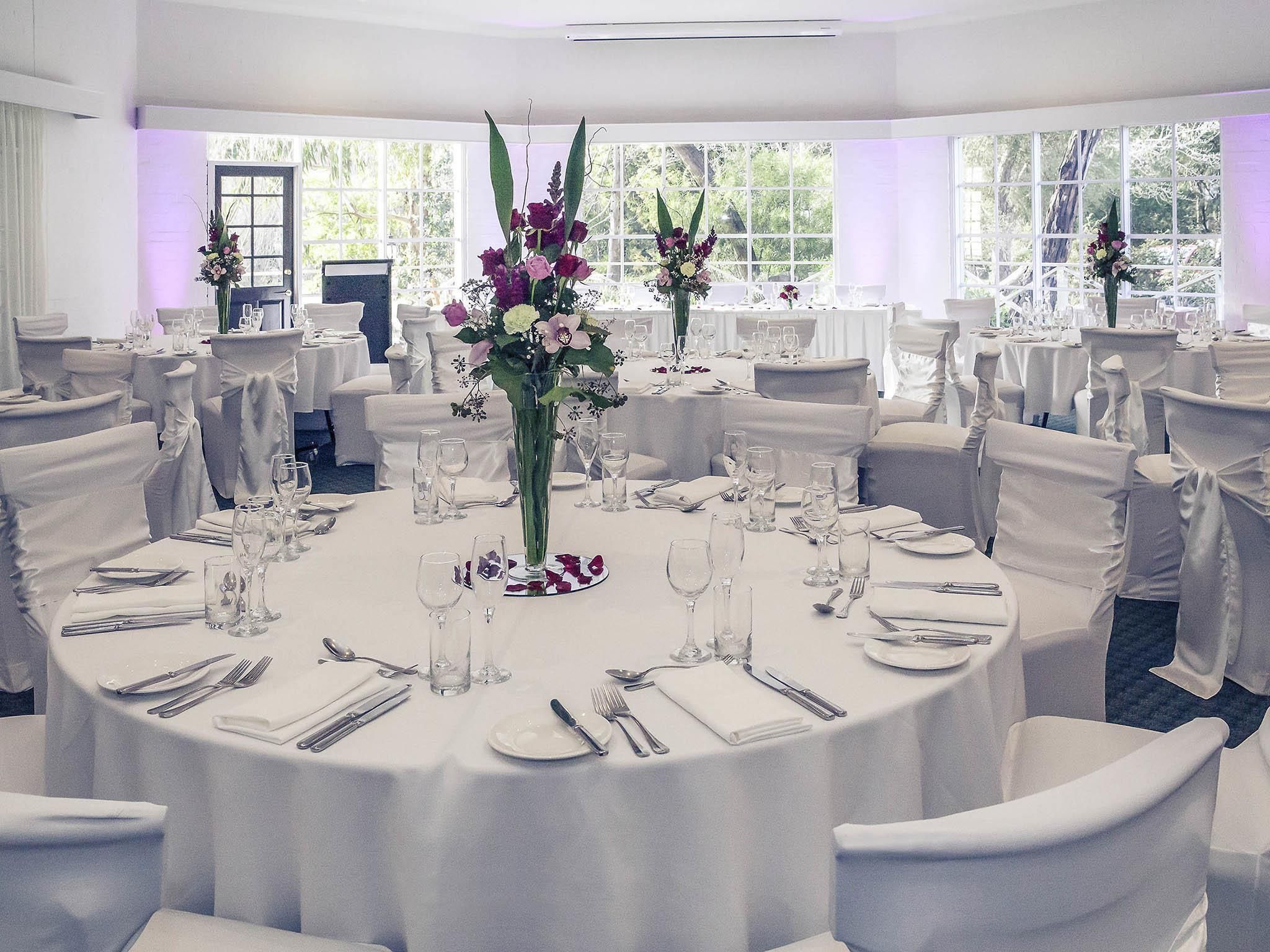 Un salón de banquetes con mesas redondas, manteles blancos y sillas con fundas blancas.