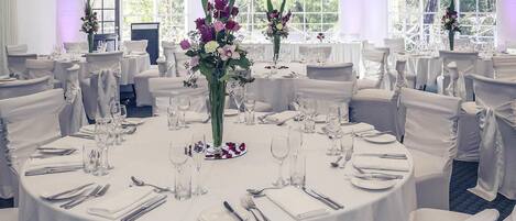 Un salón de banquetes con mesas redondas, manteles blancos y sillas con fundas blancas.