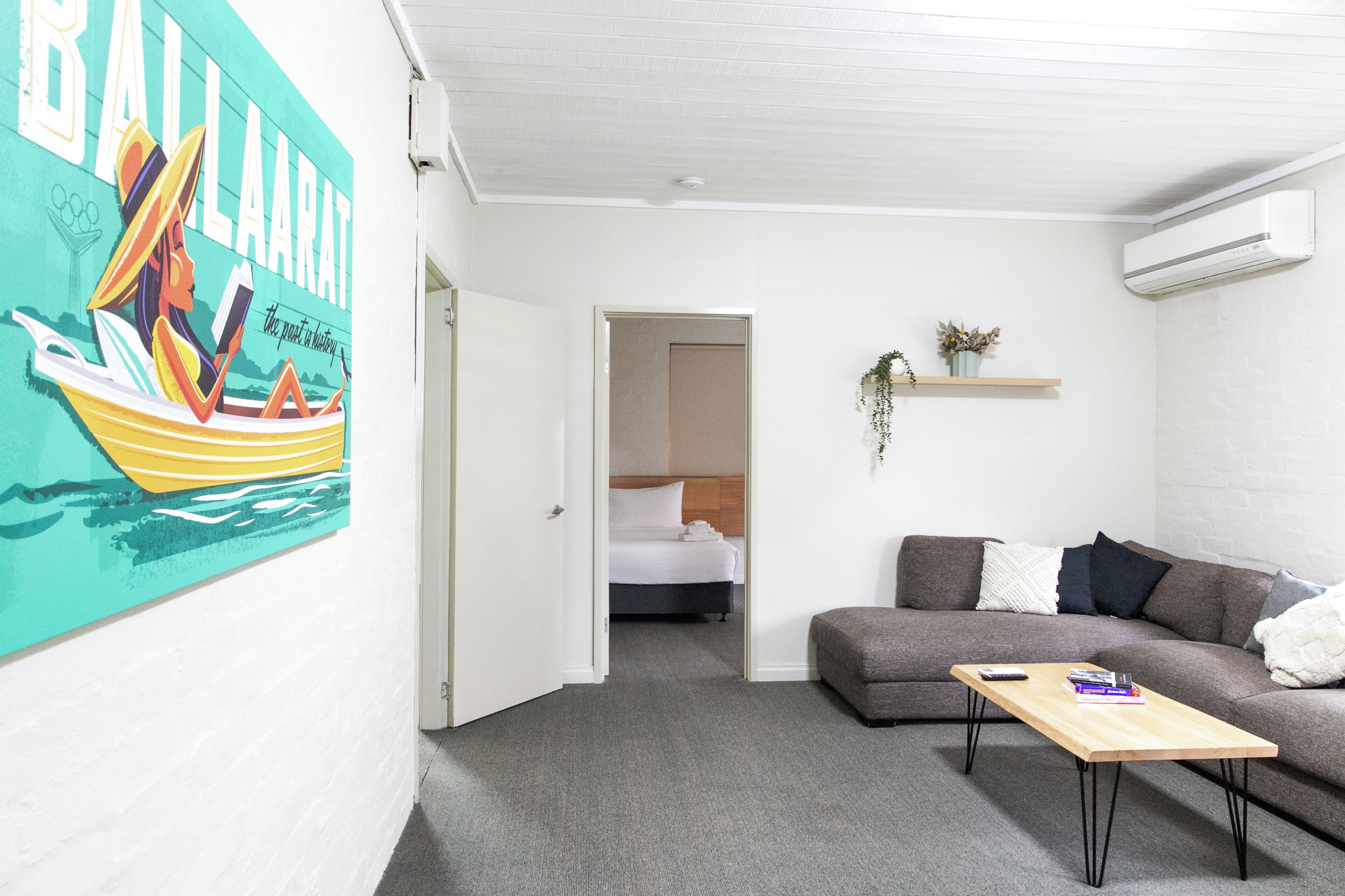 A modern hotel room with a bed, a sofa, a coffee table, a wall-mounted air conditioner, and a painting of a boat.
