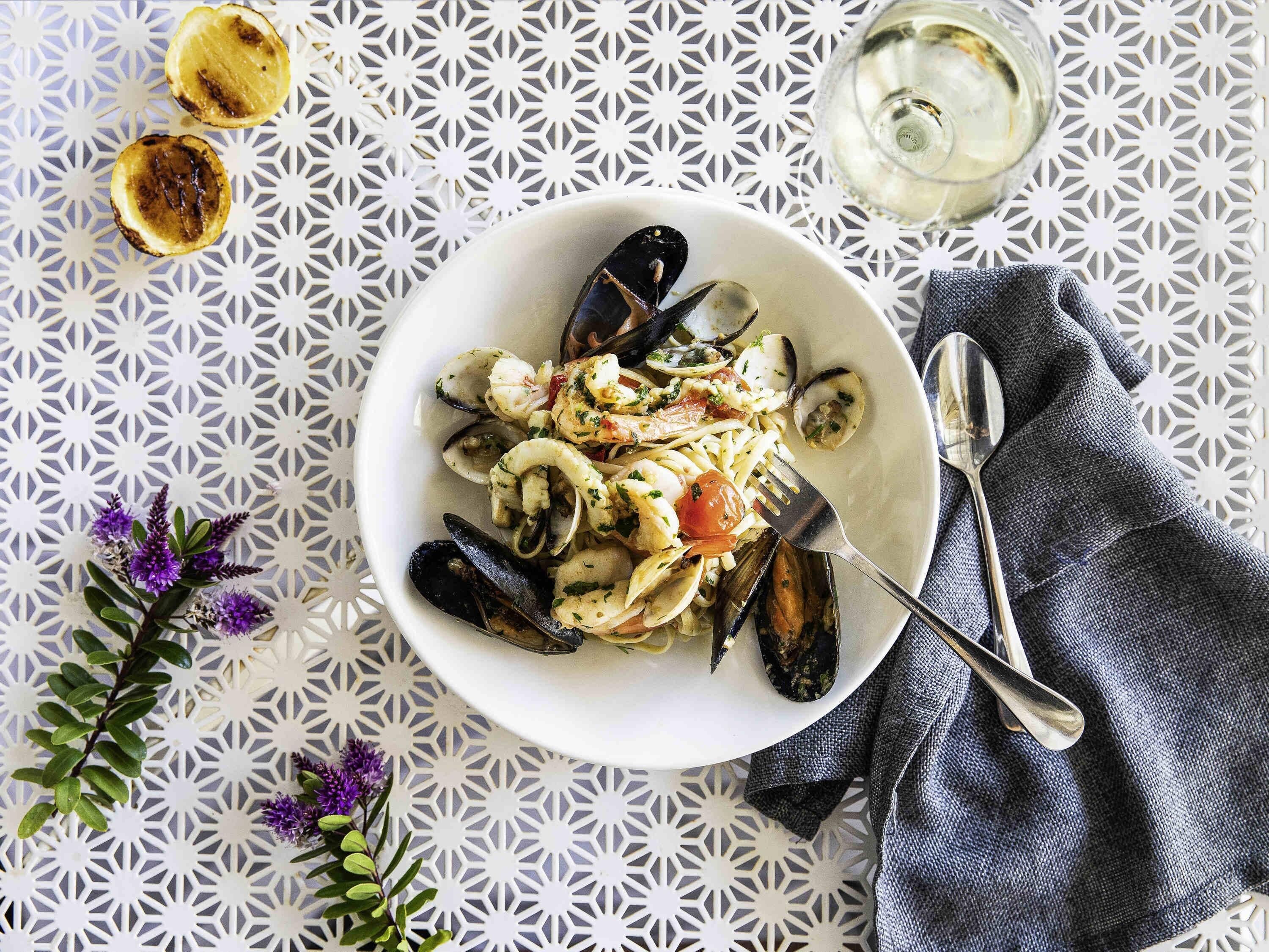 A plate of seafood pasta with mussels, shrimp, and clams, accompanied by a glass of white wine and a fork on a patterned tablecloth.