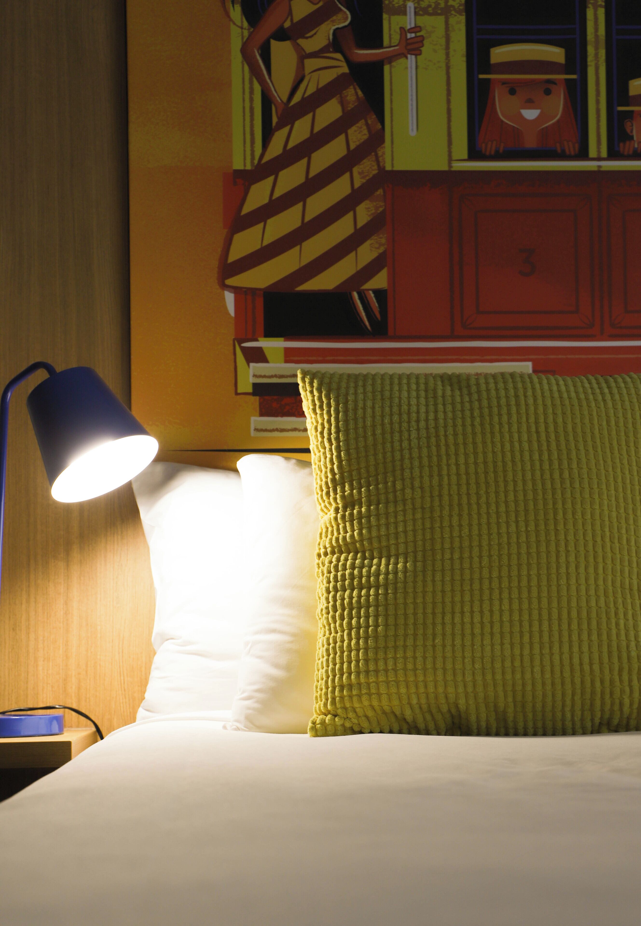 A bed with a white pillow and a textured yellow pillow, a blue lamp, and a wall with a cartoon character.