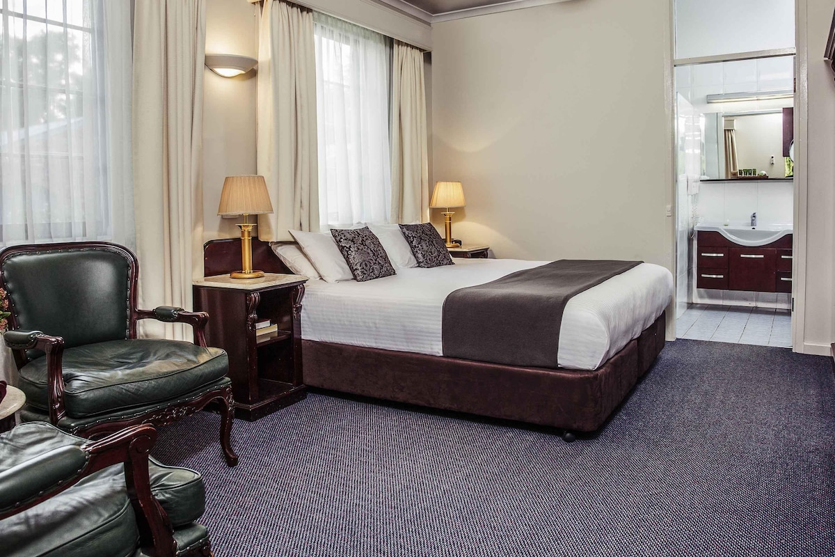 Une chambre d’hôtel avec un grand lit, un bureau, une chaise, une coiffeuse avec un miroir et une fenêtre avec des rideaux.