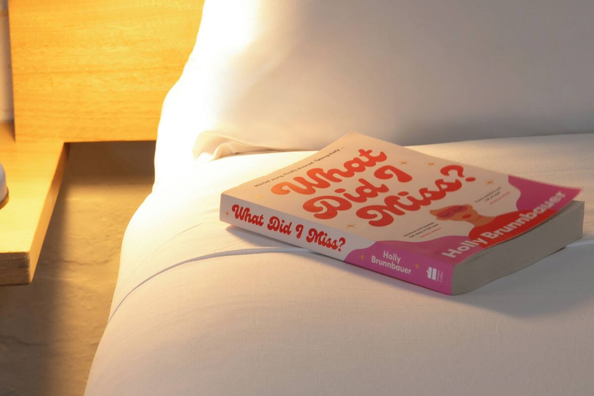 Een boek met de titel 'What Did I Miss?' op een bed, met een wit kussen en een lamp op een nachtkastje.