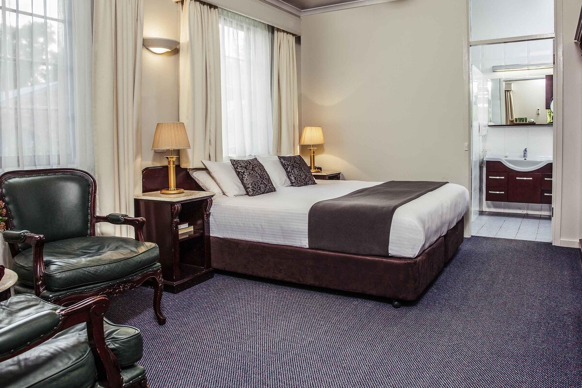 A hotel room with a large bed, a desk, a chair, a vanity with a mirror, and a window with curtains.