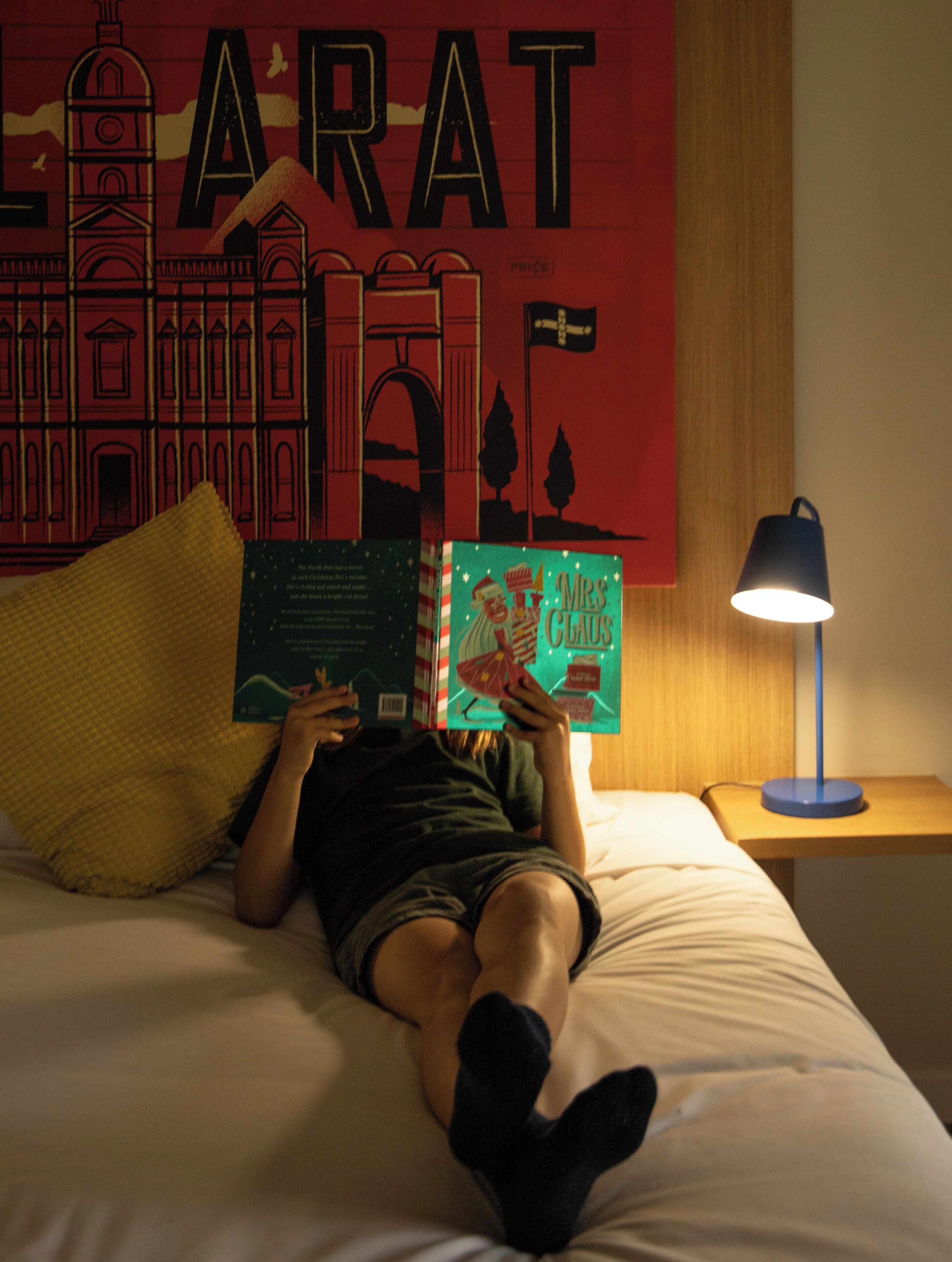 A person lying on a bed reading a book, with a lamp on a nightstand beside them.