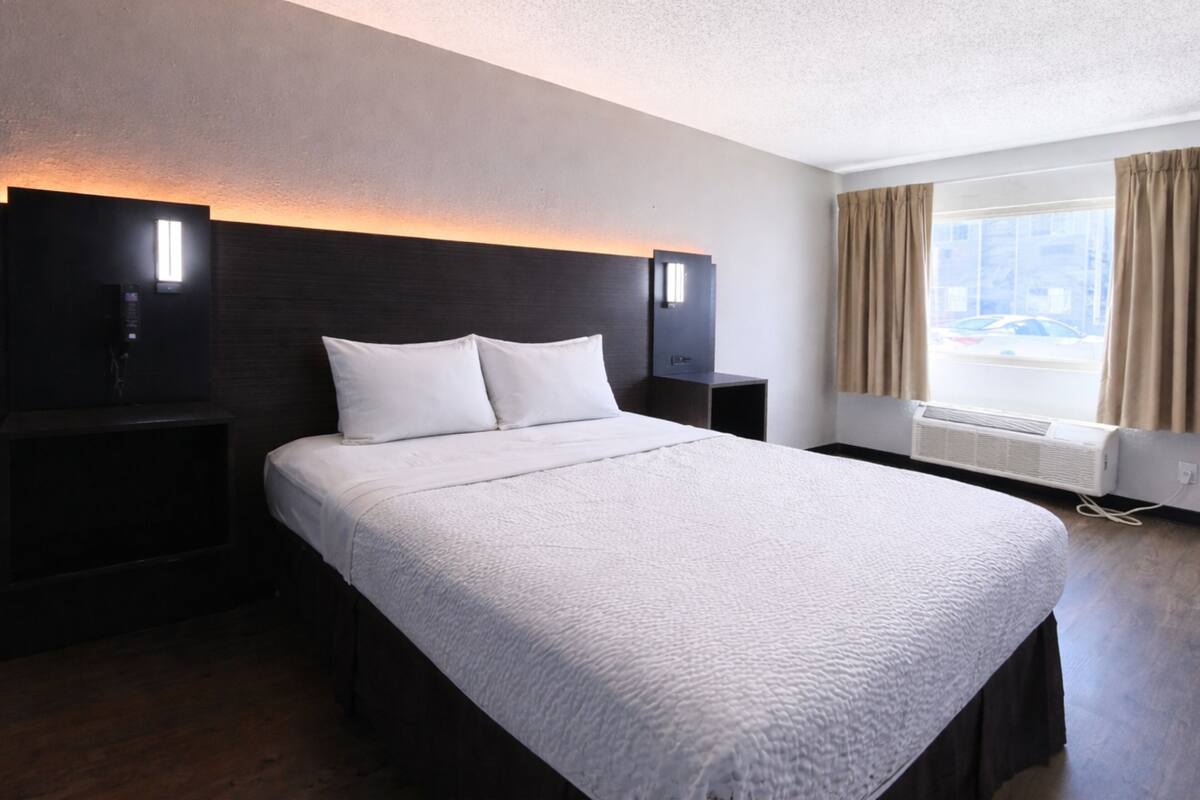 A hotel room with a large bed, two bedside tables, a nightstand, a wall-mounted lamp, a window with curtains, and an air conditioning unit.