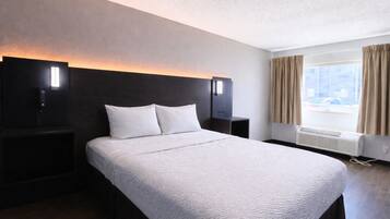 A hotel room with a large bed, two bedside tables, a nightstand, a wall-mounted lamp, a window with curtains, and an air conditioning unit.
