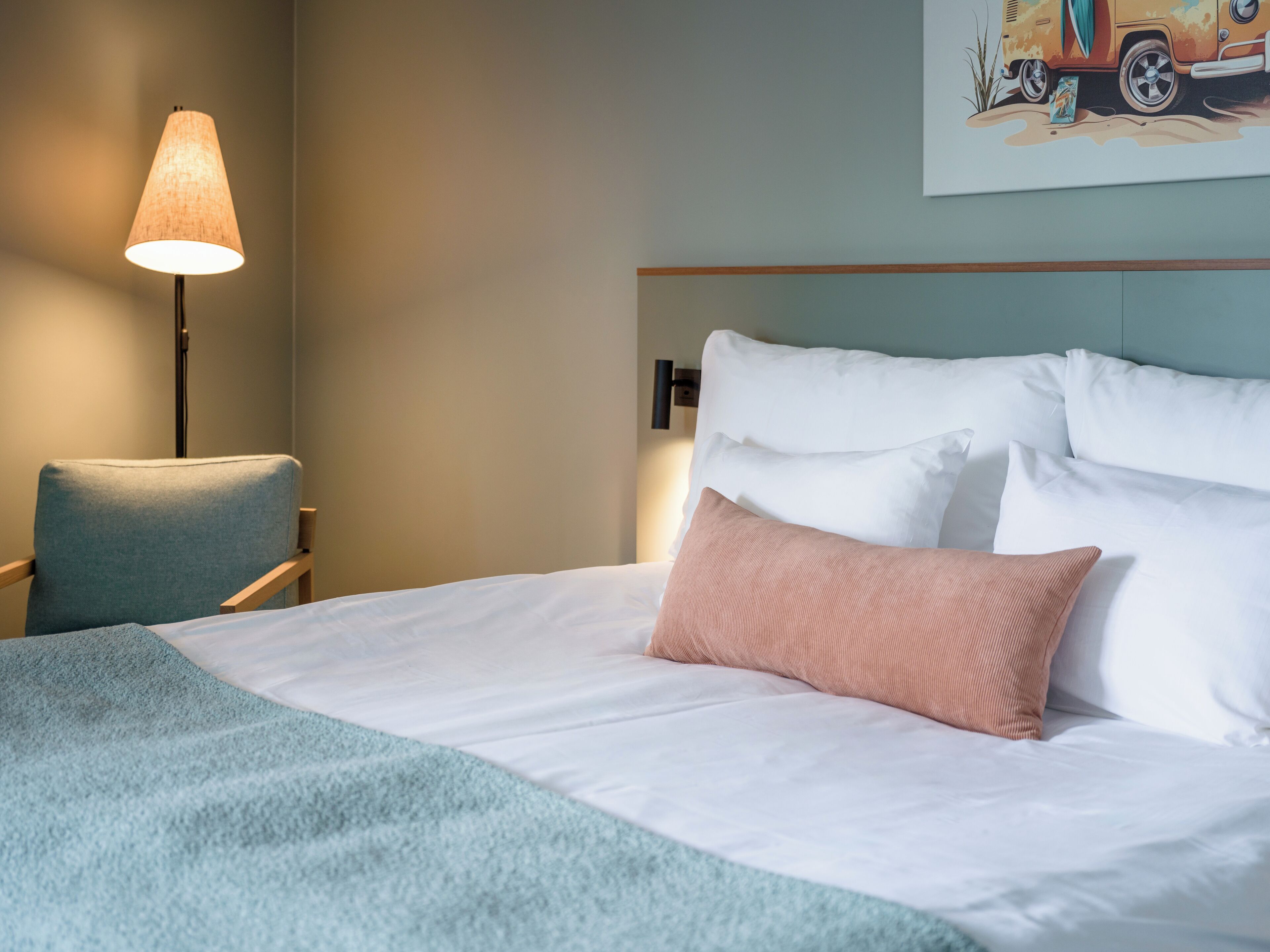 A neatly made bed with white and pink pillows, a blue chair, and a lamp.