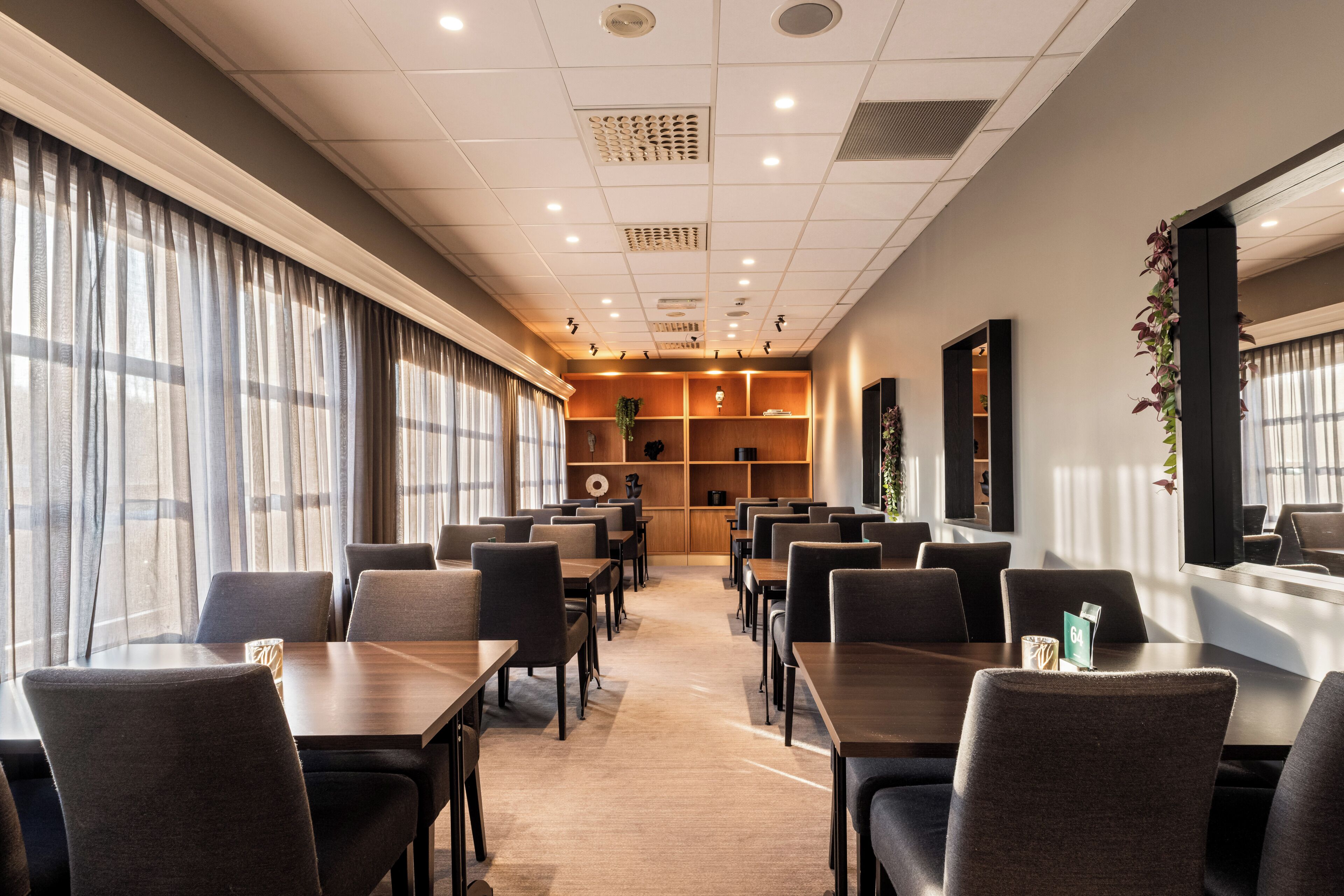 A hotel dining area with tables and chairs, a wooden reception desk, and a large window with curtains.