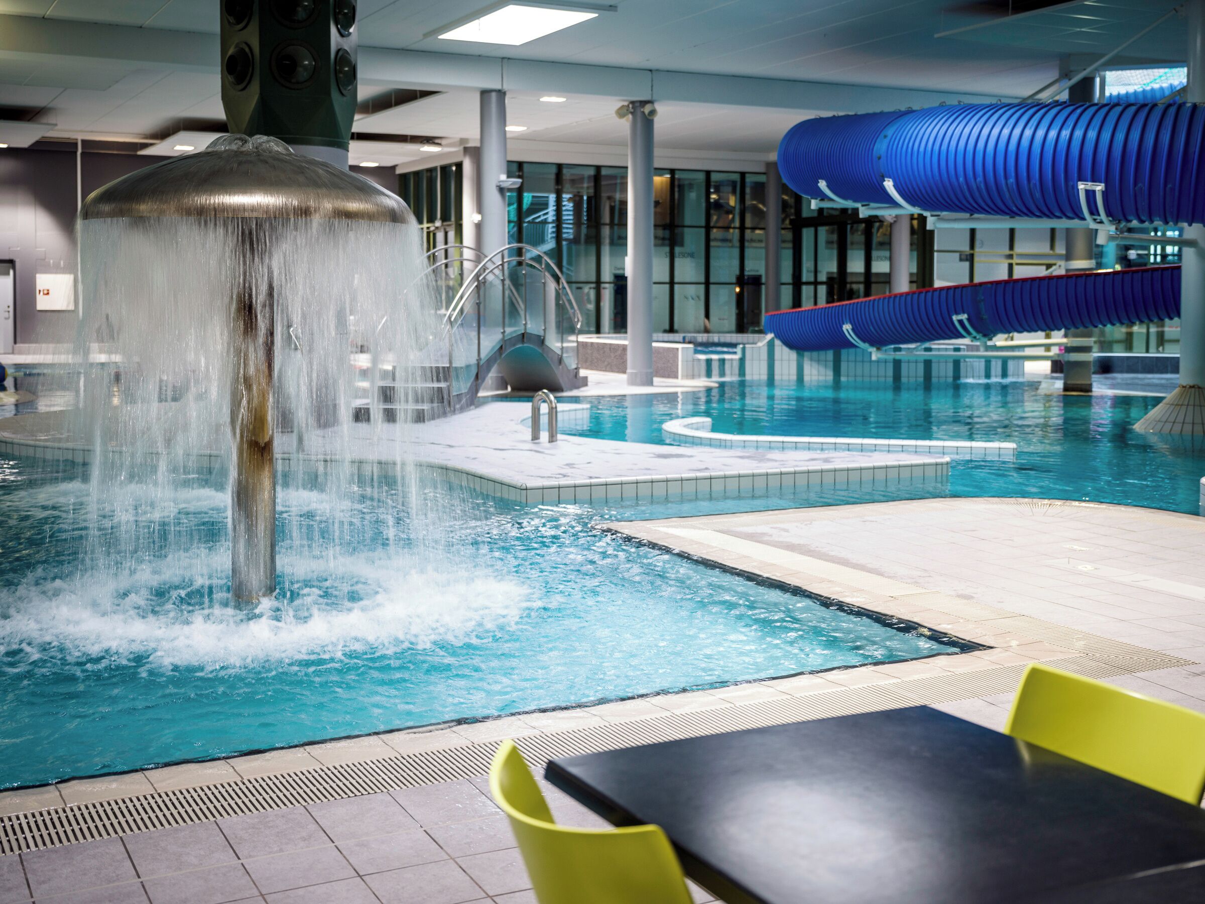 An indoor swimming pool with a water fountain, a slide, and seating areas.