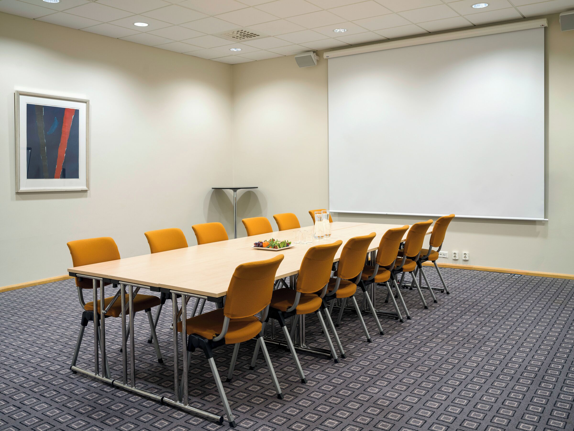 Un salón de conferencias con una larga mesa, sillas anaranjadas, una pantalla de proyector y un cuadro en la pared.