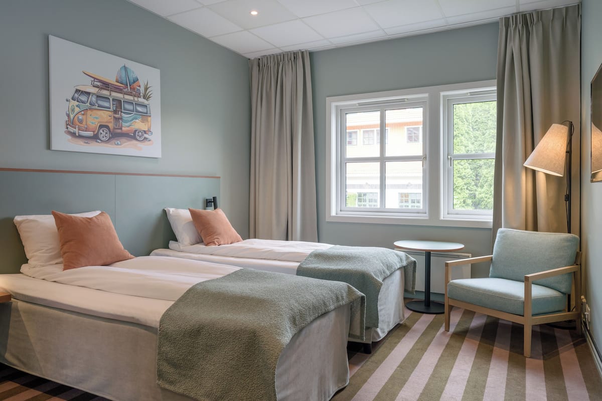 Habitación de hotel con dos camas, una mesita redonda pequeña, una silla, una ventana con cortinas y un cuadro con motivos playeros en la pared.