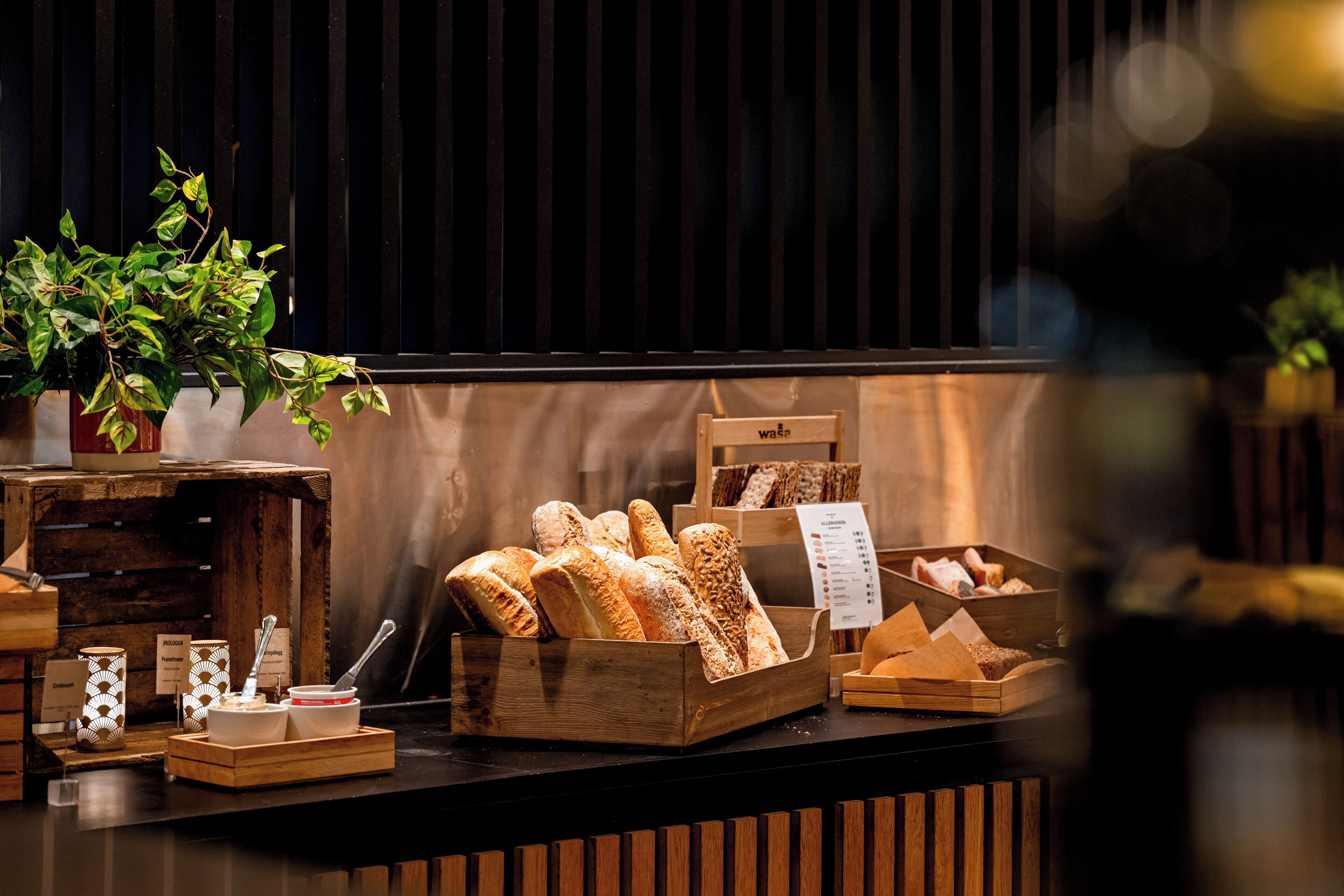 A bakery display with various breads and pastries, wooden crates, and a potted plant.