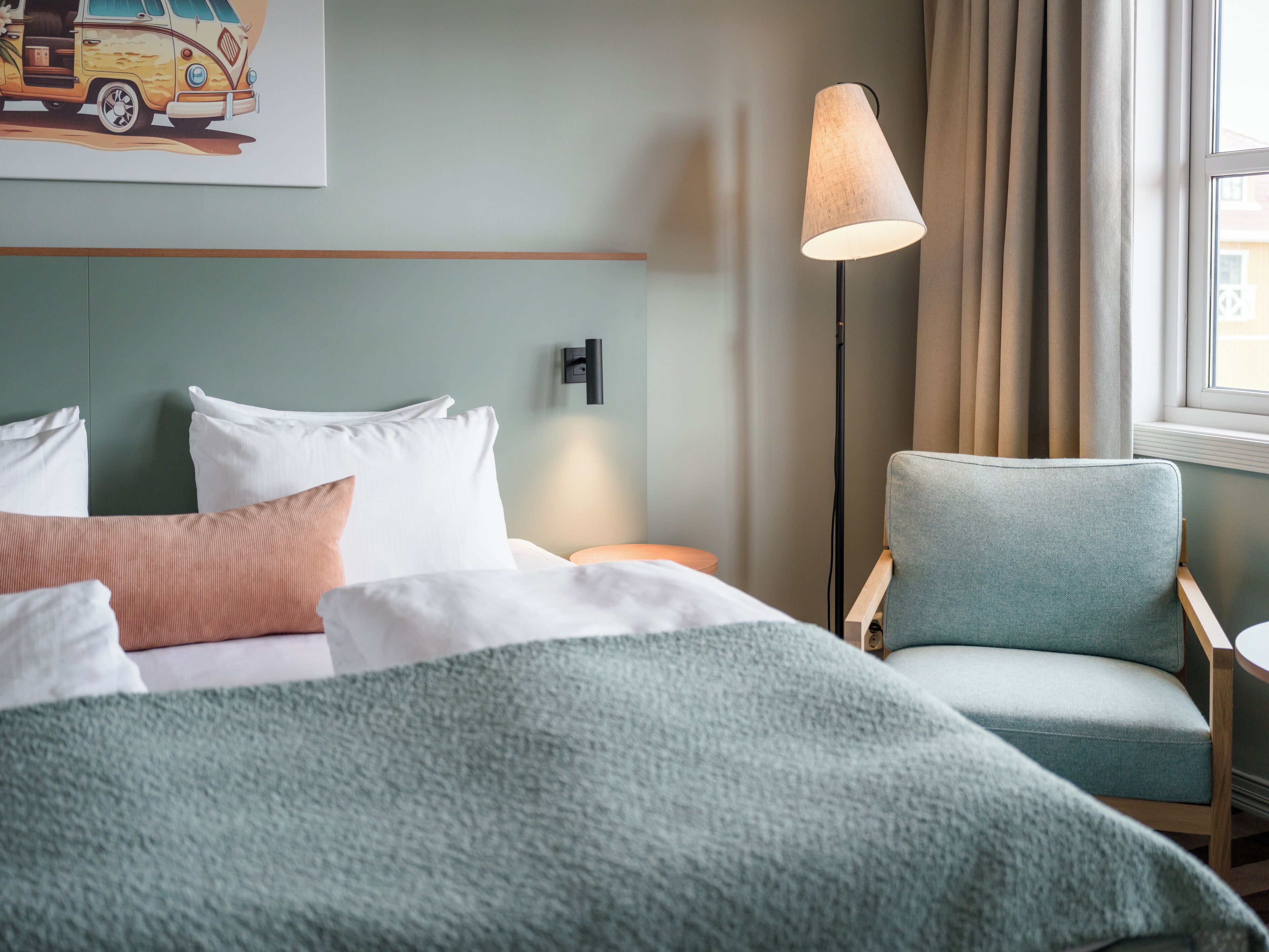 A hotel room with a bed, a chair, a lamp, and a painting on the wall.