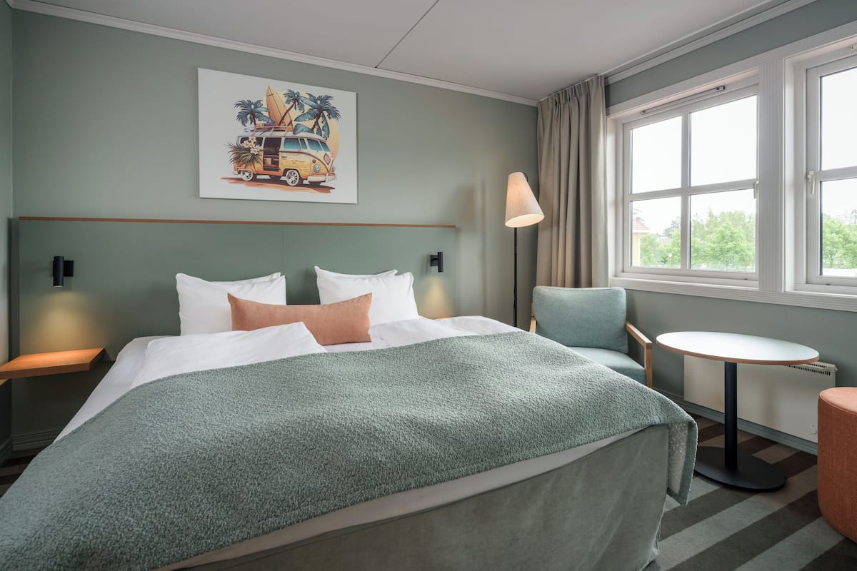 Una habitación de hotel con una cama, una silla, una mesita, una lámpara y una ventana con cortinas.