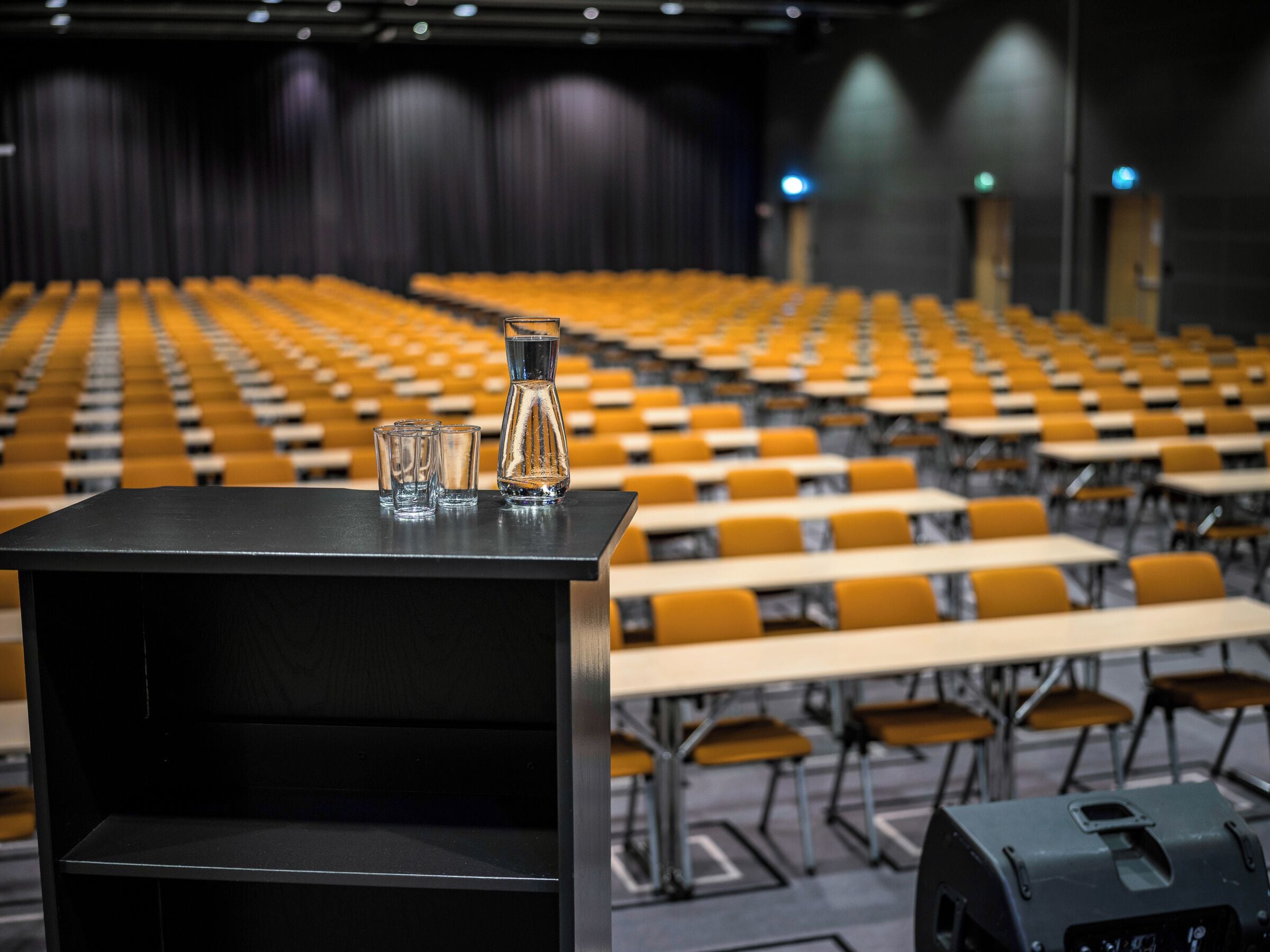 An auditorium with rows of yellow chairs and tables, a podium with a microphone, and a projector on a stand.