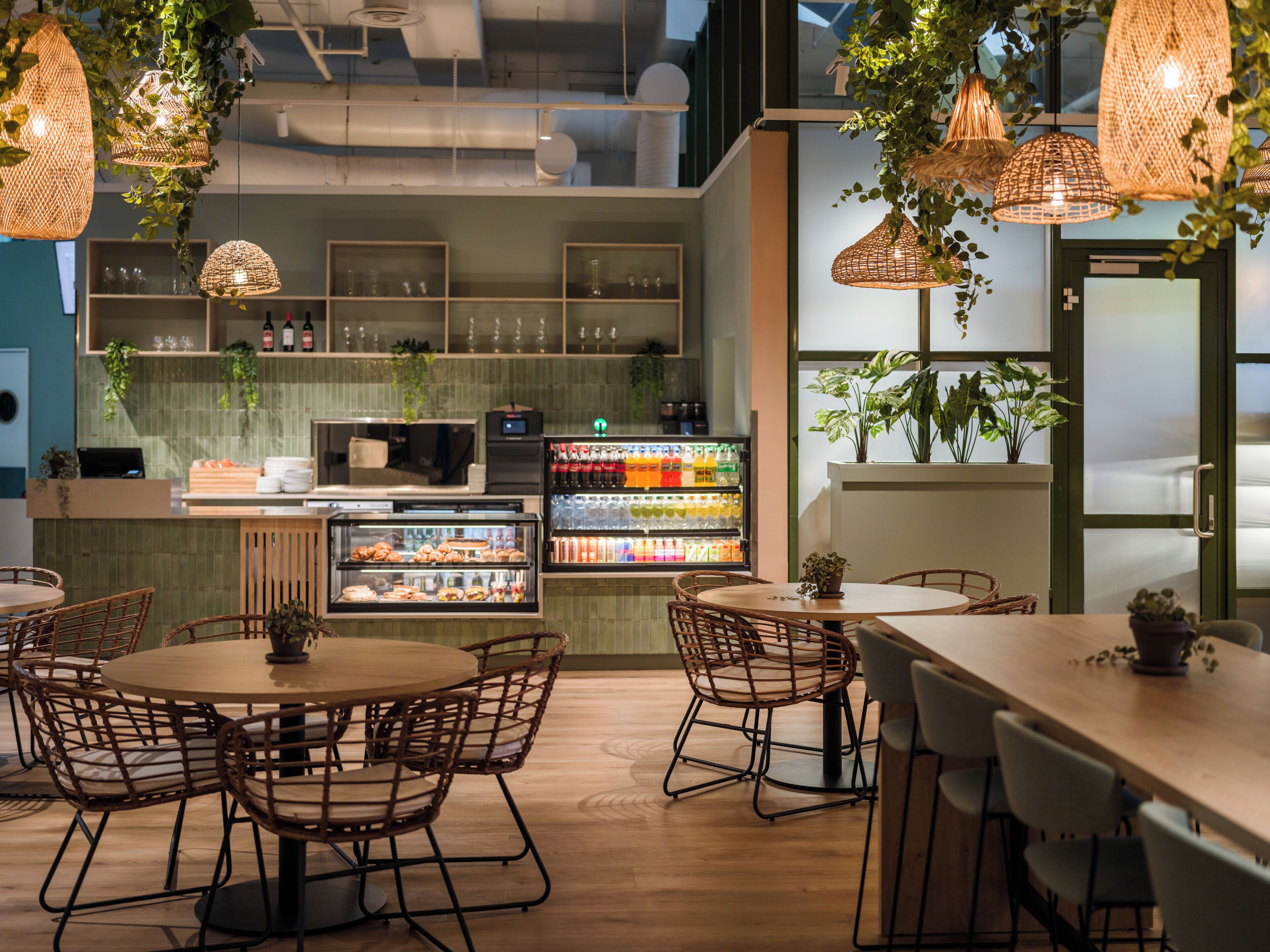 A cafe interior with round tables and wicker chairs, a refrigerated display case with drinks, and hanging wicker lamps.