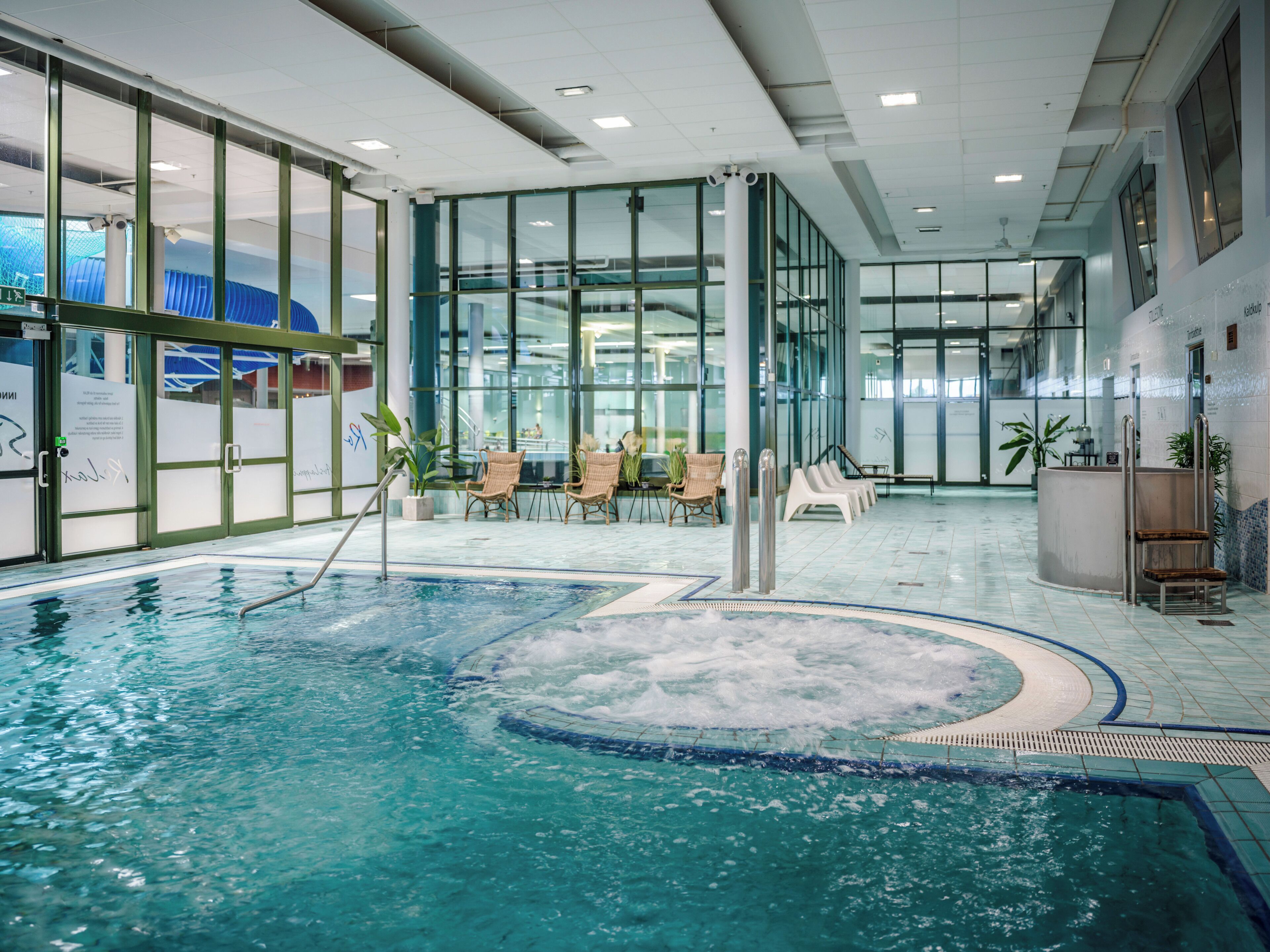 Una piscina interior con agua cristalina, rodeada de paredes de vidrio y equipada con sillones de mimbre.