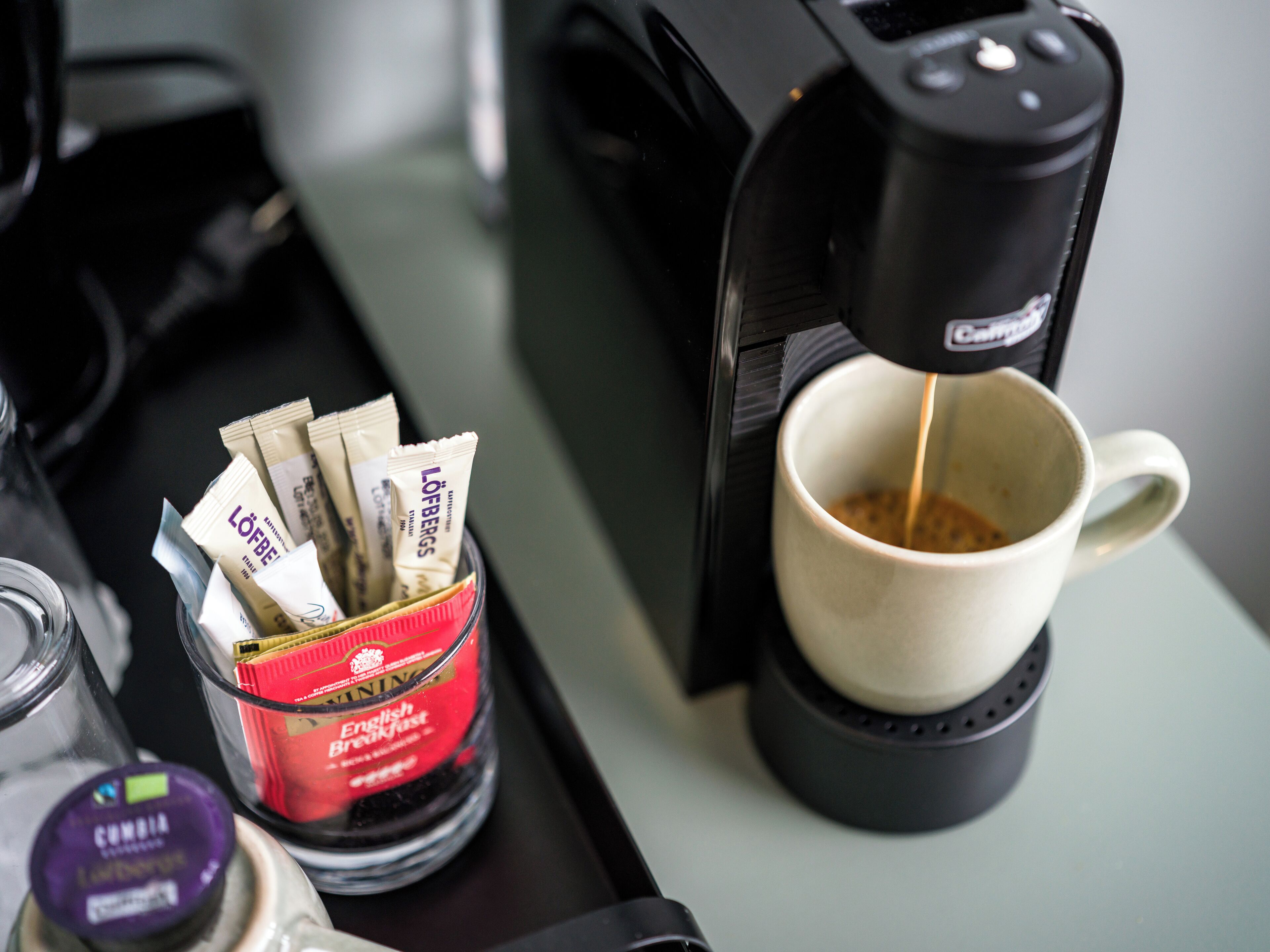 Una cafetera sirviendo café en una taza, un recipiente de vidrio con cápsulas de café y un tarro de crema.