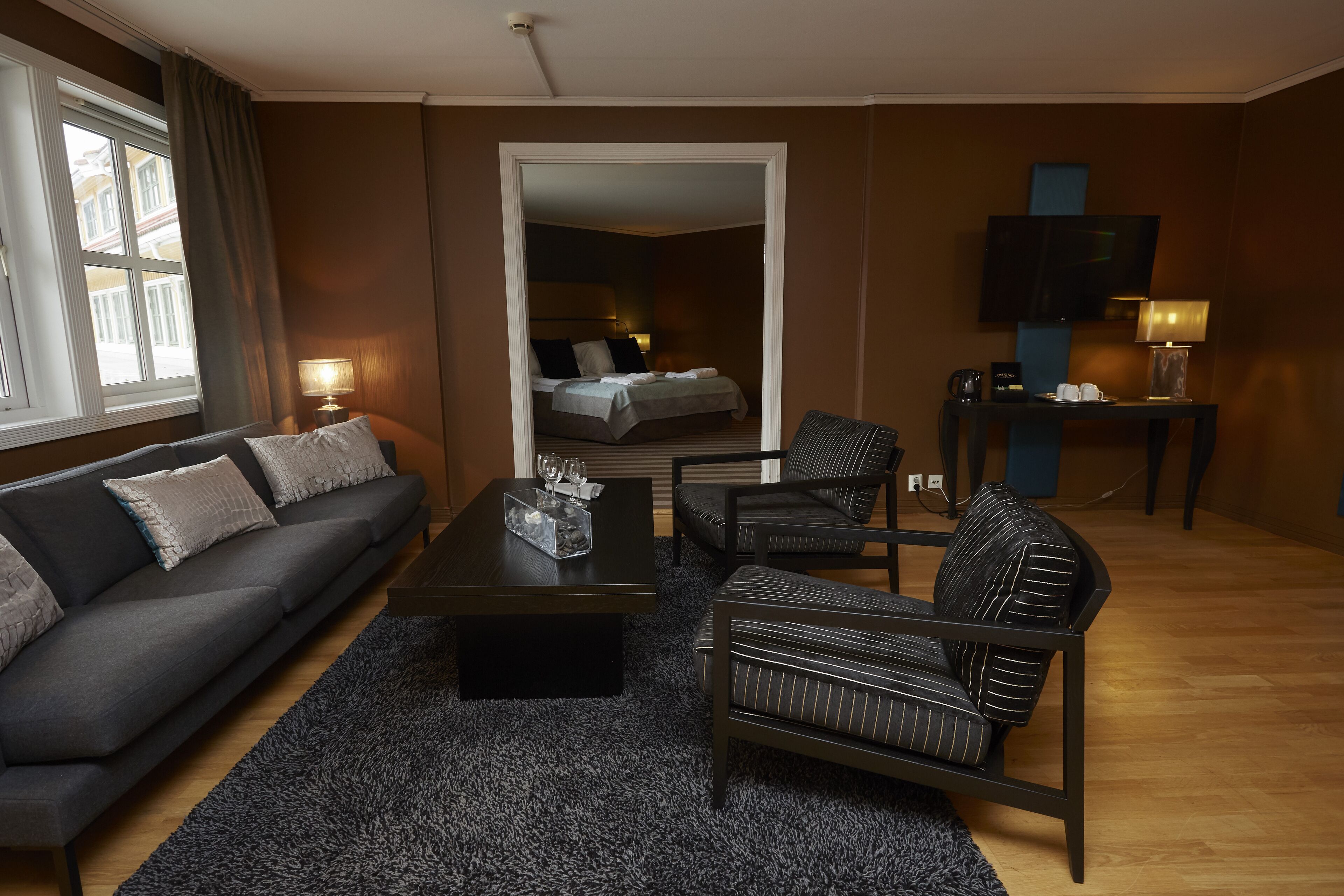 Una habitación de hotel moderna con una pared marrón oscuro, un sofá, un sillón a rayas, una mesita de café y un televisor montado en la pared.