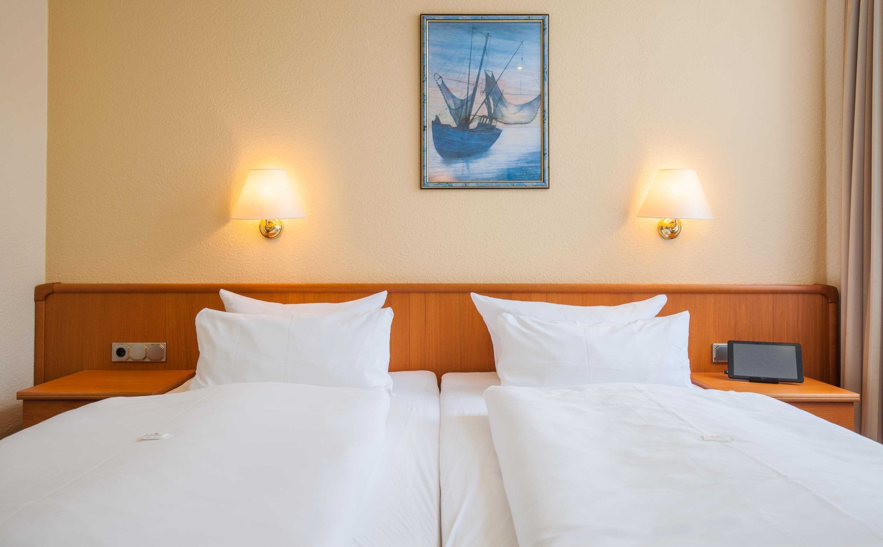A hotel room with a double bed, two bedside tables, a nightstand, a lamp, a framed picture on the wall, and a small electronic device on the nightstand.