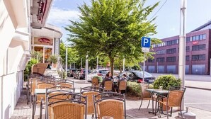 Un café al aire libre con sillas y mesas de mimbre, un árbol y un edificio con un letrero de estacionamiento 'P' al fondo.