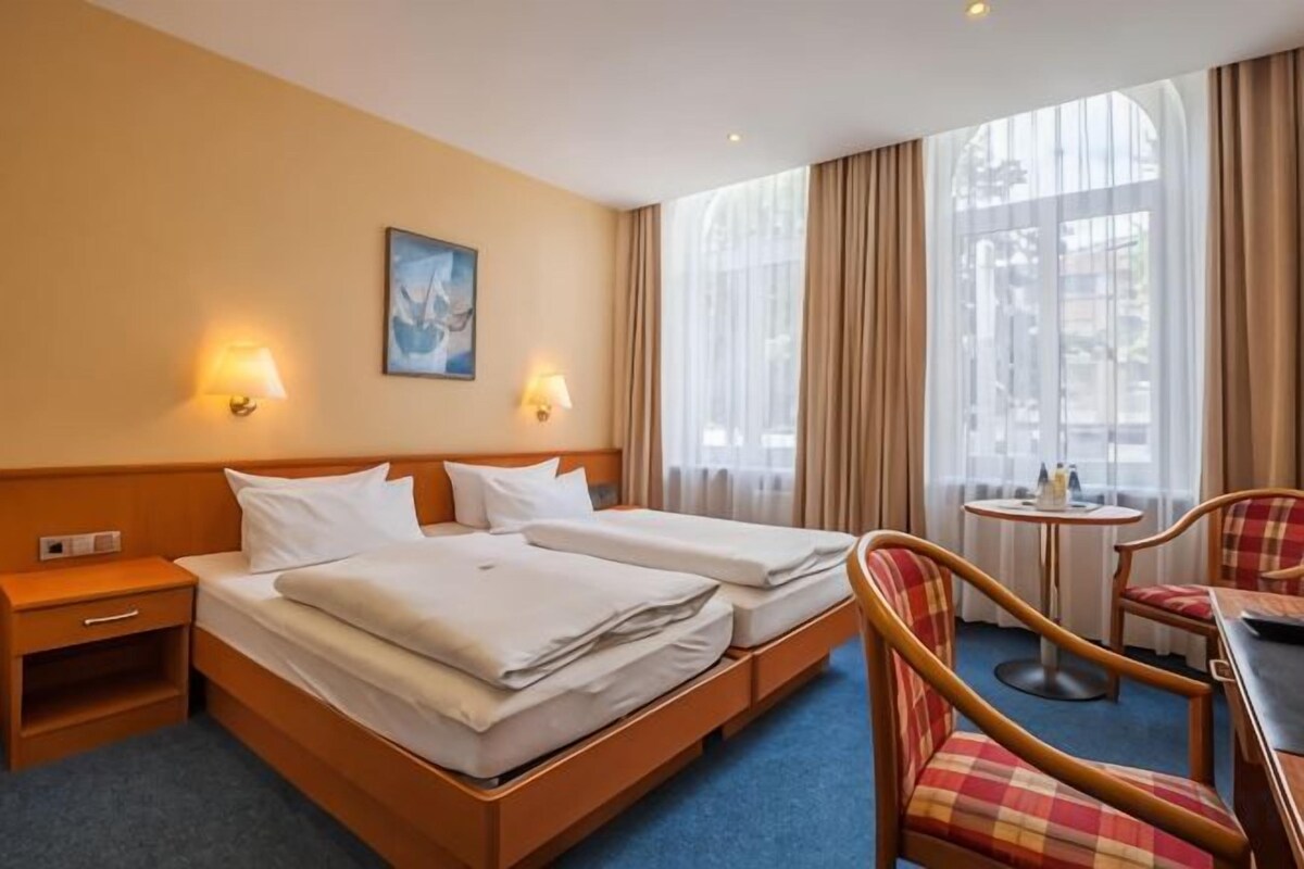 Une chambre d’hôtel avec deux lits, une petite table, une chaise et une fenêtre avec des rideaux.