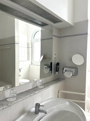 Une salle de bain avec un grand miroir, un lavabo avec un robinet, un distributeur de serviettes et un verre d’eau posé sur le comptoir.