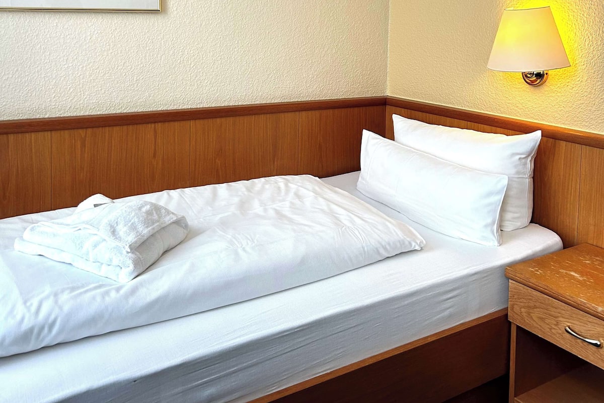 Una cama bien tendida con sábanas y almohadas blancas, un cabecero de madera, una mesita de noche con lámpara y un mueble auxiliar de madera.