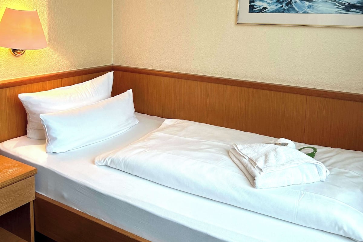 Una cama bien hecha con sábanas blancas, un cabecero de madera y una mesita de noche con una lámpara.