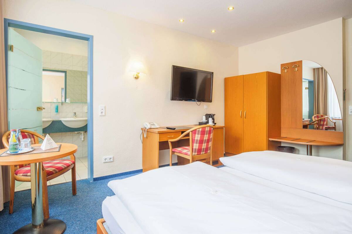 Habitación de hotel con cama, escritorio con silla, televisor y baño visible a través de una puerta abierta.