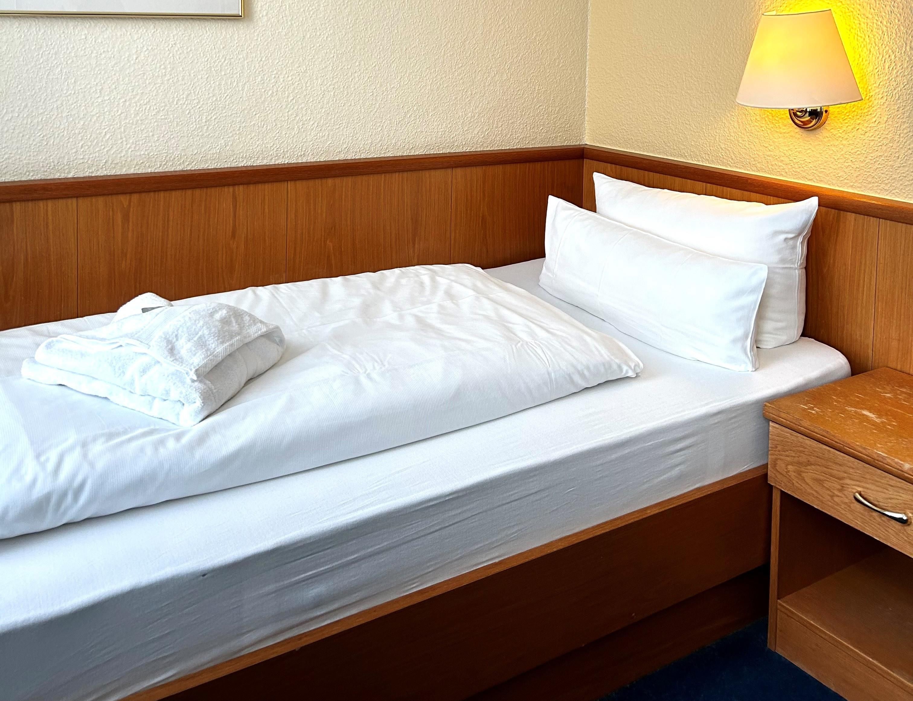 A neatly made bed with white linens and pillows, a wooden headboard, a bedside table with a lamp, and a wooden nightstand.
