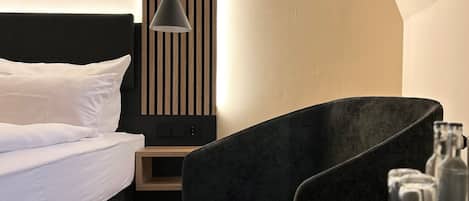A modern hotel room with a bed, a chair, a small table with glasses, and a wall-mounted lamp.