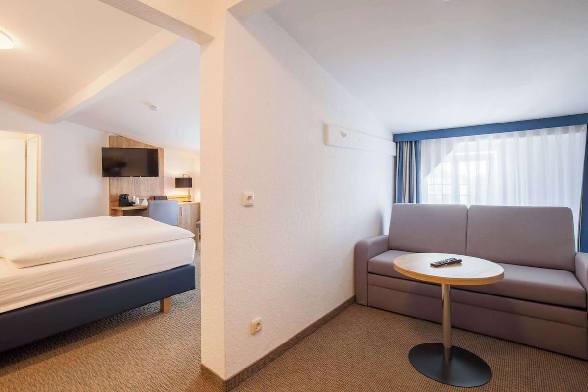 Habitación de hotel con cama, sofá, una mesita, televisor y ventana con cortinas.