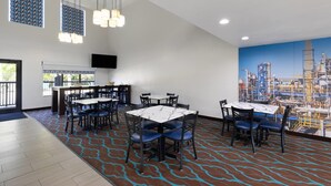 A modern dining area with multiple tables and chairs, a large mural of an industrial scene, and a flat-screen TV mounted on the wall. - Baymont by Wyndham Moss Point/Pascagoula (Moss Point)