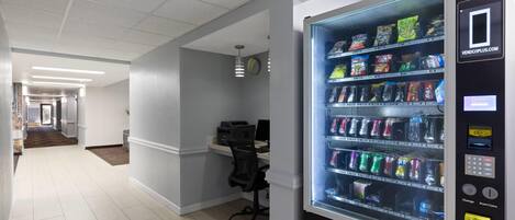 A vending machine with a variety of snacks and drinks, located in a hallway with a desk and chair.