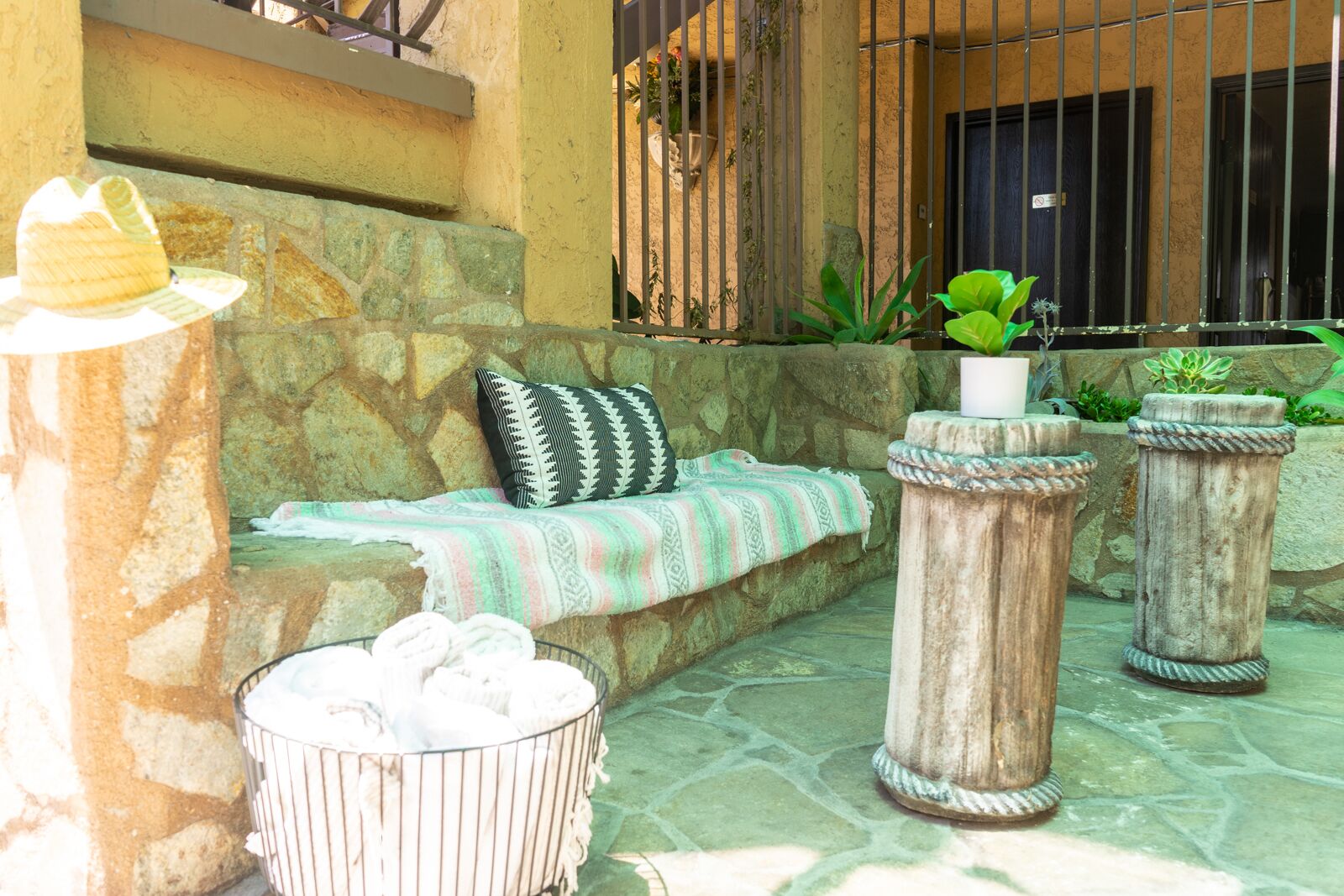 A stone bench with a striped blanket, a metal trash bin, and two stone planters with plants.