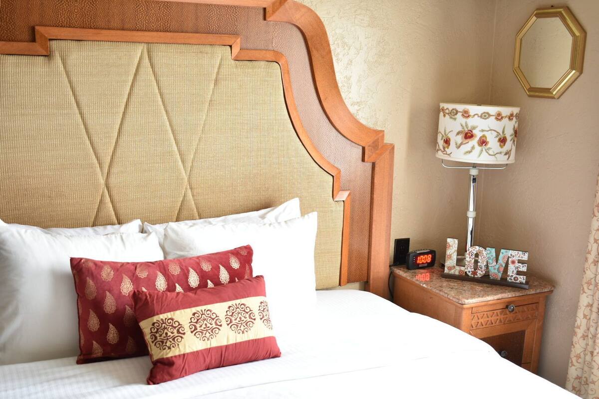 A neatly made bed with white and red pillows, a wooden headboard, a bedside table with a lamp and a clock, and a decorative sign saying "LOVE".