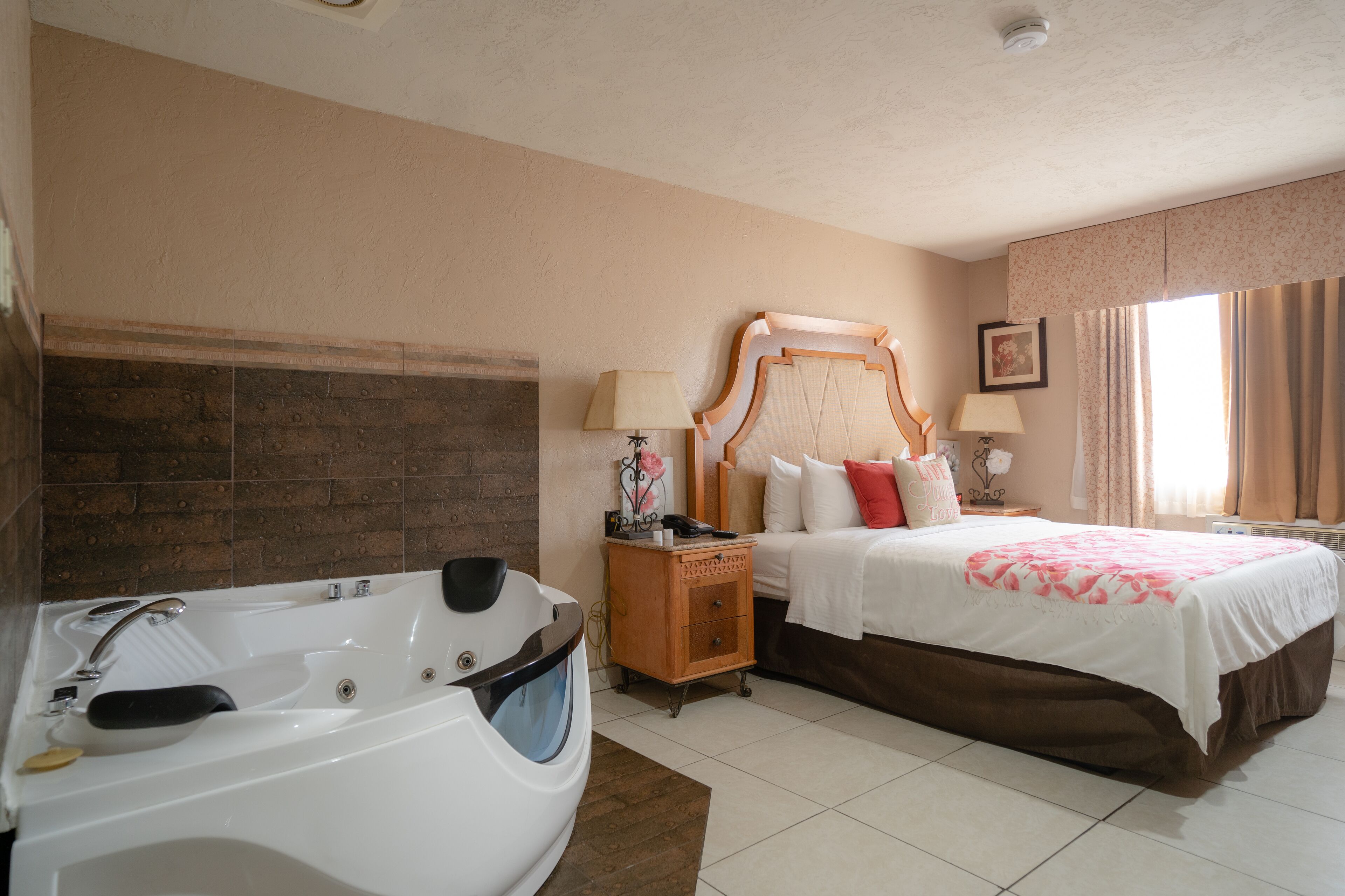 A hotel room with a large bathtub, a double bed with a wooden headboard, and a nightstand with a lamp.