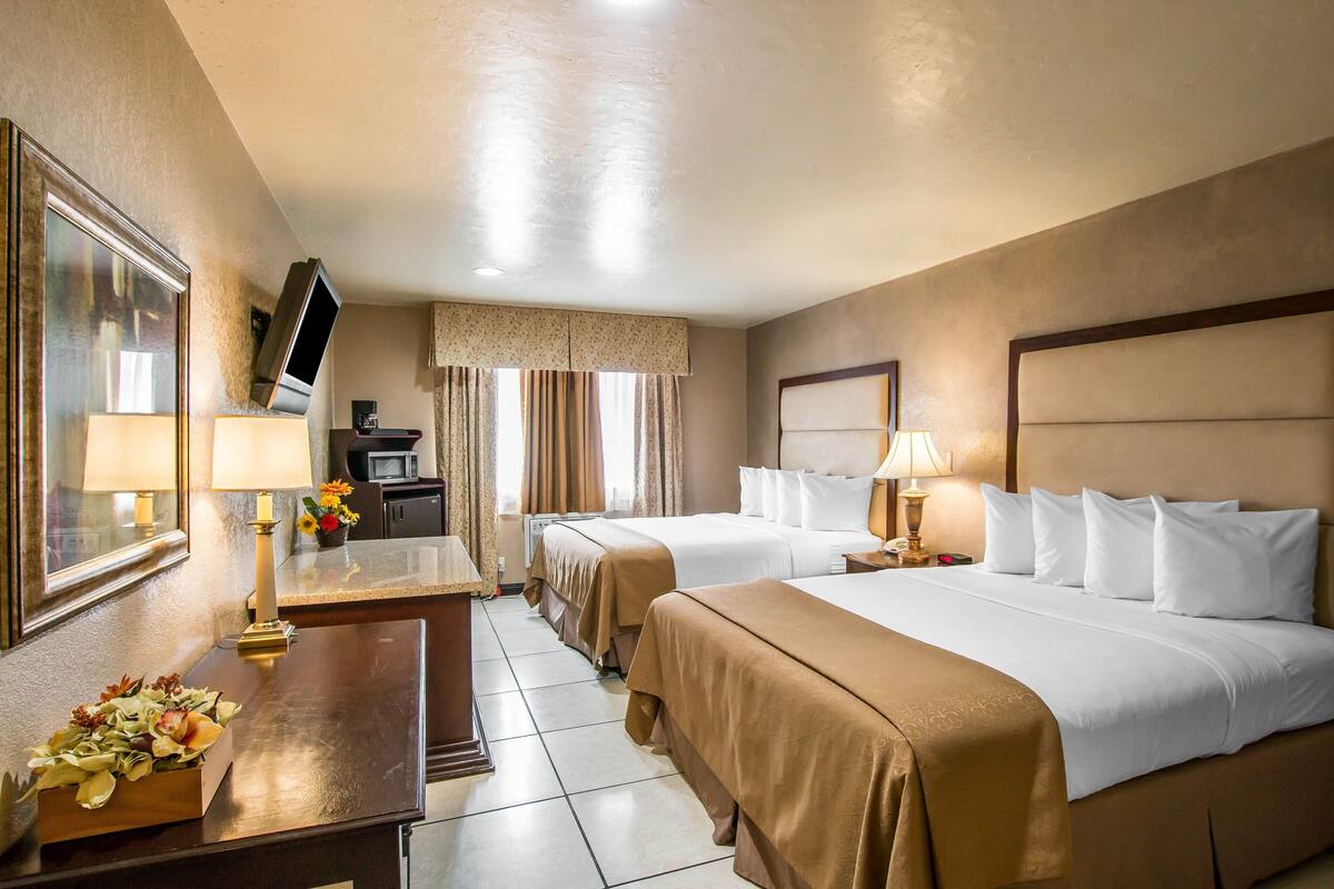 A hotel room with two beds, a desk with a lamp, a TV, and a small table with flowers.