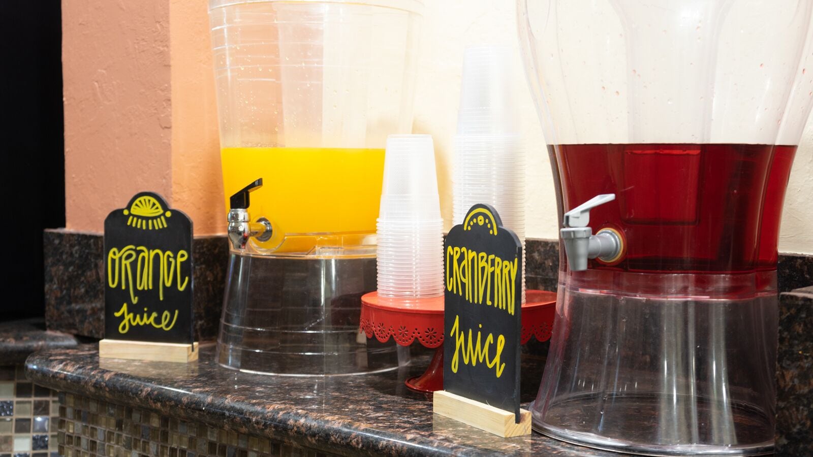 Uma estação de bebidas com dispensadores de sumo de laranja e cranberry, e uma bebida vermelha.