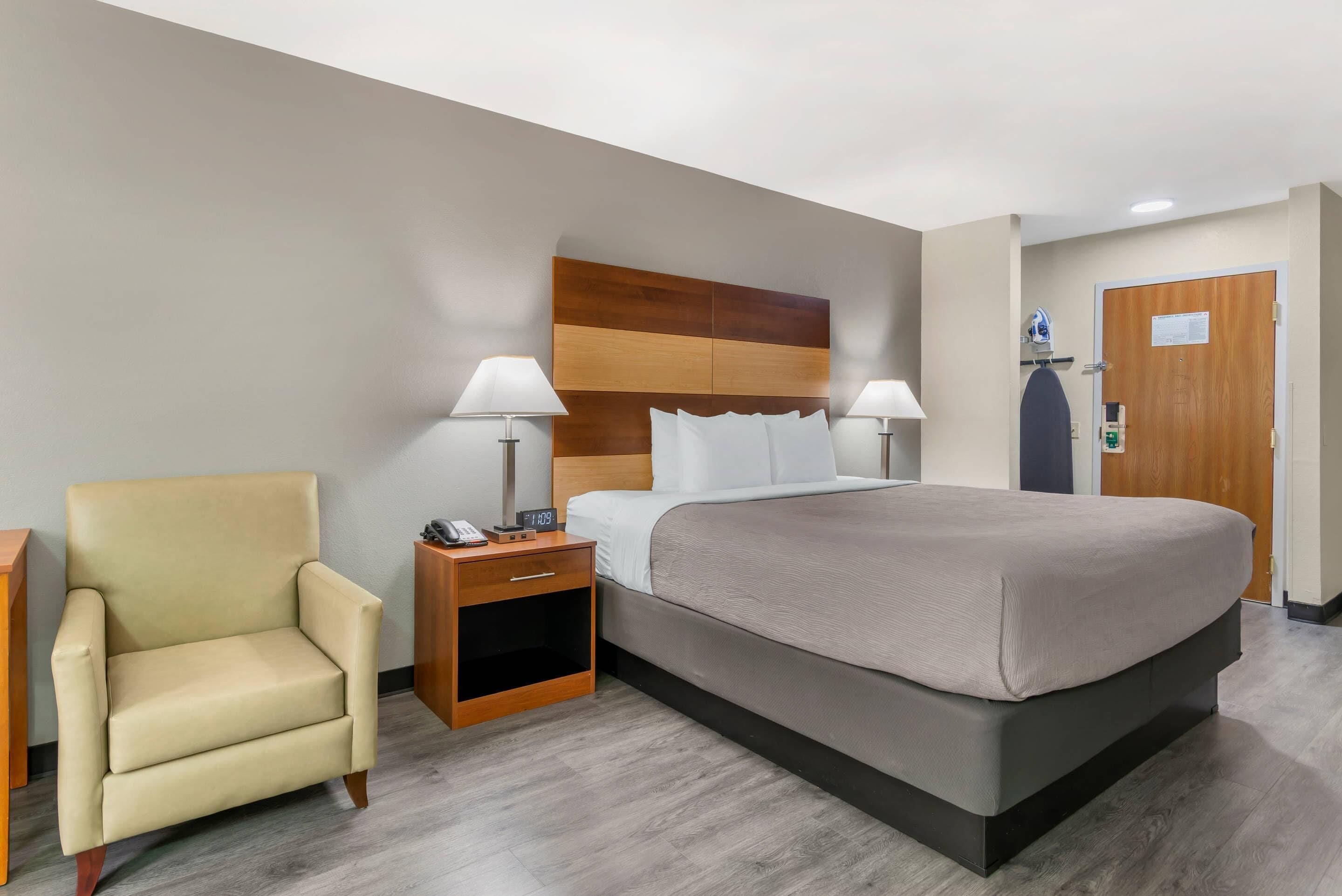 A hotel room with a large bed, a chair, two bedside tables with lamps, a wooden headboard, and a door with a coat hanging on it.