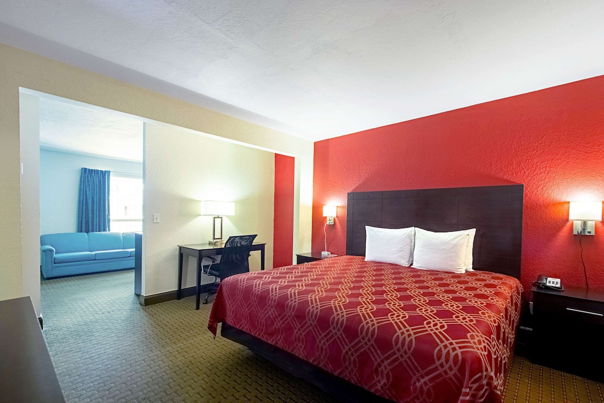 Habitación de hotel con una decoración en tonos rojo y beige, una cama grande con almohadas blancas, un escritorio con una silla y un sofá azul en la habitación contigua.