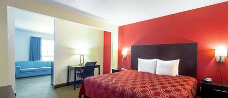 Une chambre d’hôtel aux couleurs rouge et beige, avec un grand lit doté d’oreillers blancs, un bureau avec une chaise et un canapé bleu dans la pièce adjacente.