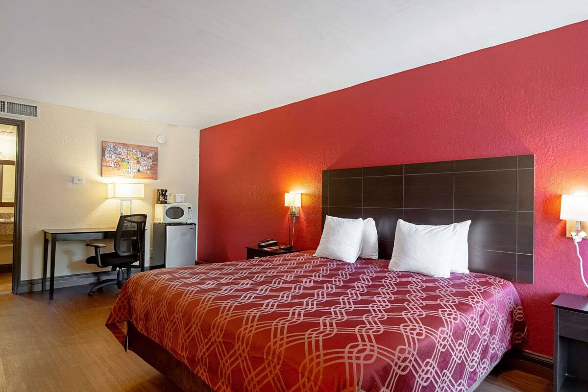 Quarto de hotel com uma cama grande, escrivaninha, cadeira, luminária e televisão fixada na parede.